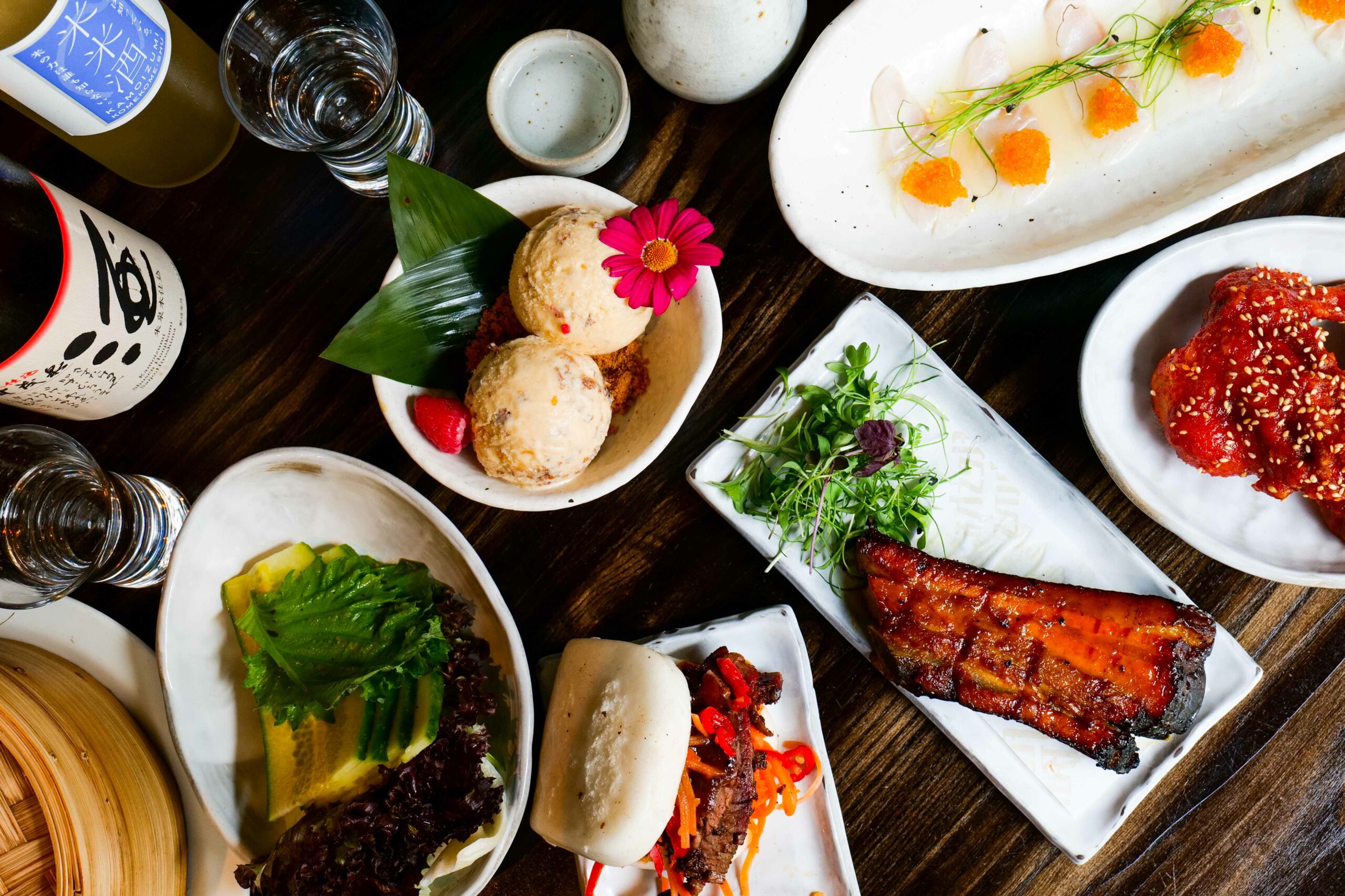 Table with various Asian dishes: sashimi, bao bun, glazed meat, ice cream, and sake.