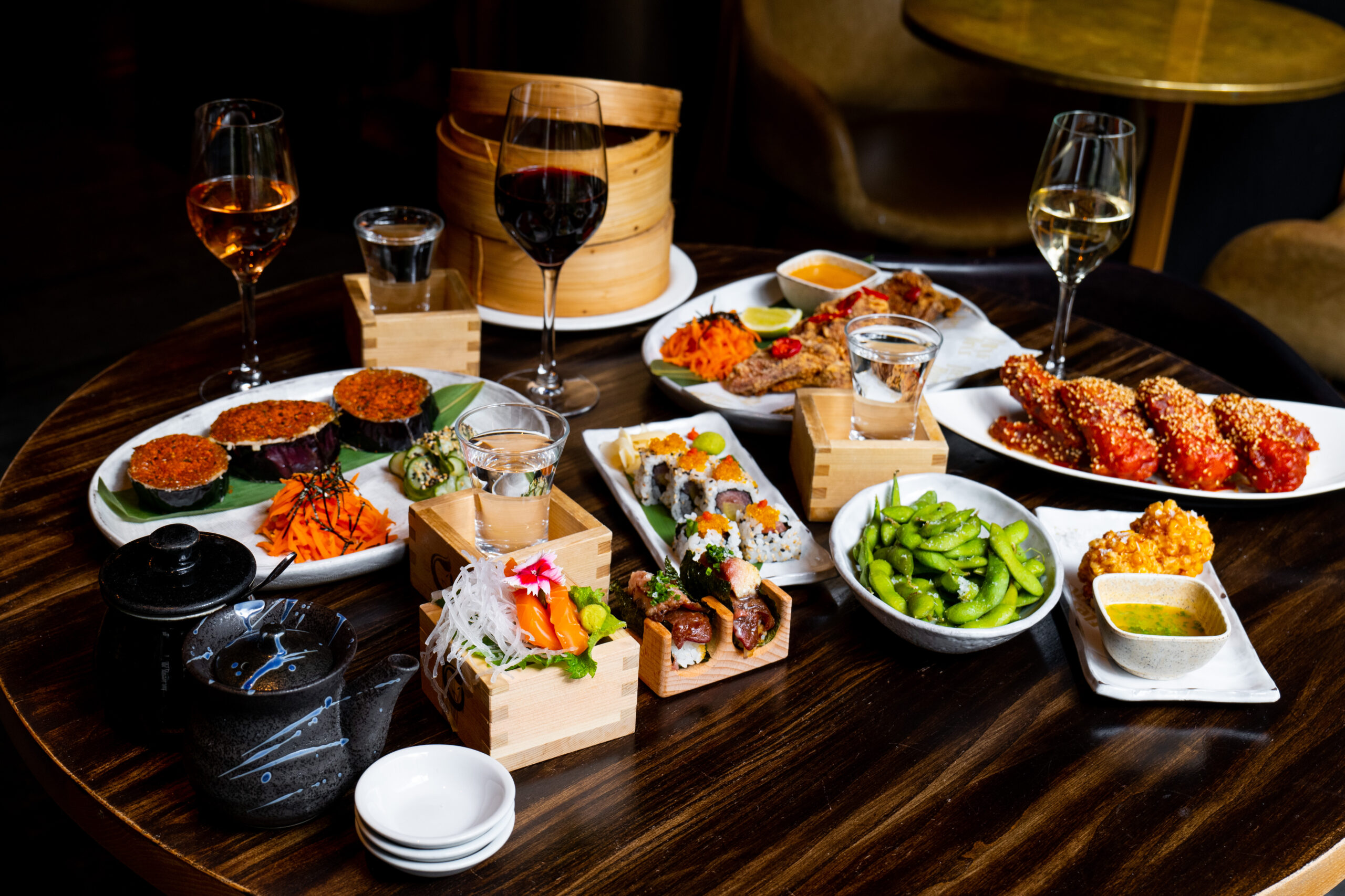 Assorted Japanese food on a wooden table, including sushi, fried chicken, edamame, and sake, with wine glasses.