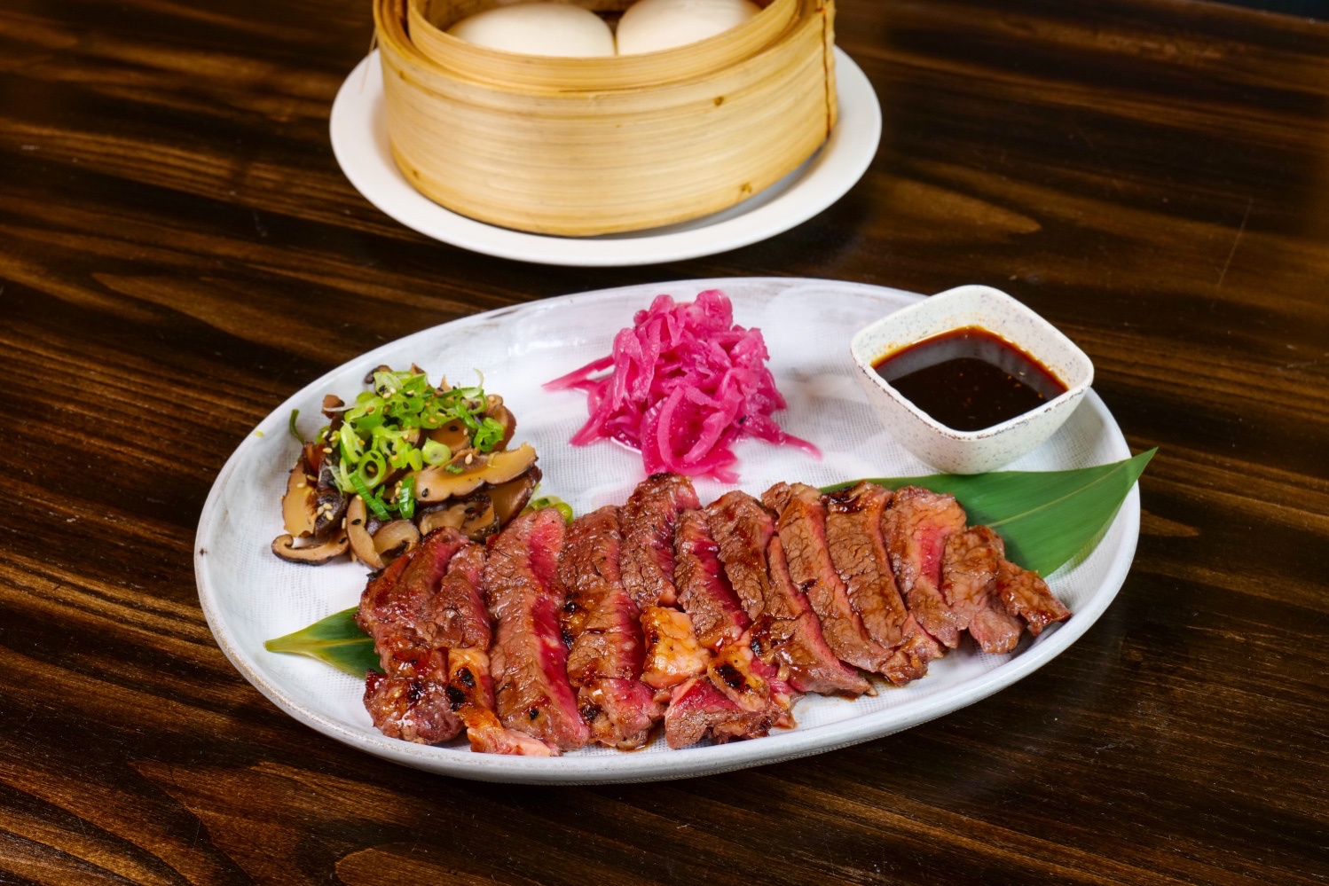 Sliced steak on a plate with mushrooms, pickled onions, and sauce. Bamboo steamer with buns in background.