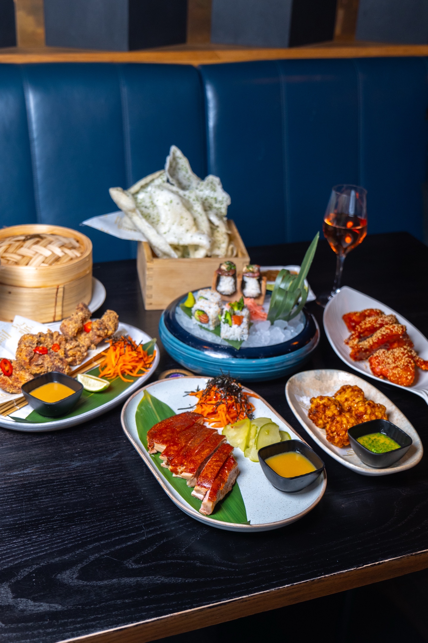 Assorted Asian cuisine: sushi, fried chicken, crispy duck, shrimp chips, and a glass of rose wine on a black table.